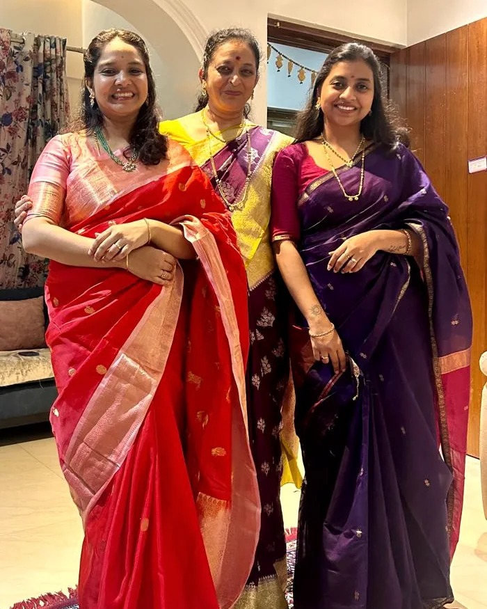 Three women of the Sangle family standing together at home in handloom silk saris: red with a gold border, yellow and purple, and deep purple
